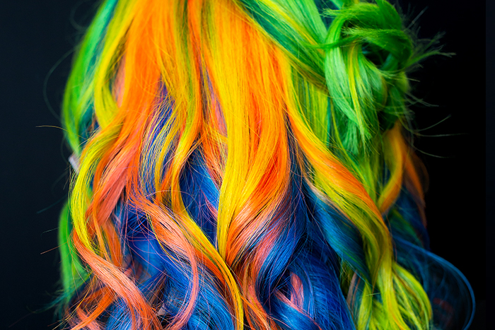 Middle aged woman with vivid rainbow hair with neon green, yellow, orange, pink, purple and deep blue sections, styled in loose curls with a small twisted bun.