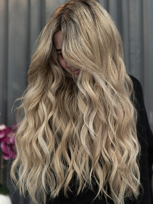 Young woman with long blonde hair with soft beige balayage style highlights, blended darker roots, and loose, textured waves with face-framing layers.