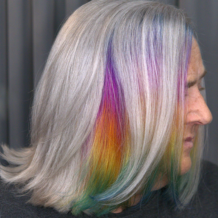 Woman with natural gray silver chin-length bob with softly flicked ends and a peekaboo rainbow prism panel blending purple, pink, orange, yellow, green and blue.