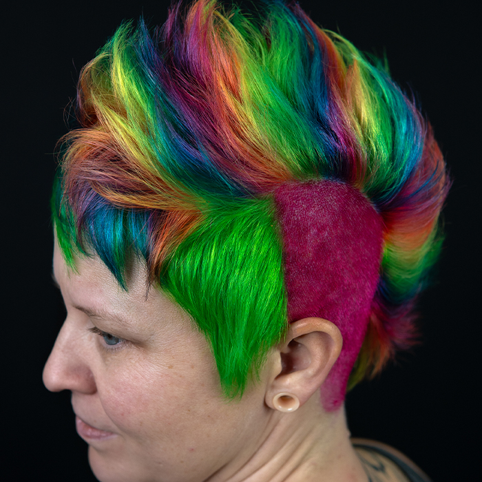 Young woman with a tall, textured rainbow mohawk, a vivid pink shaved side, and a bright green block of hair framing her face.