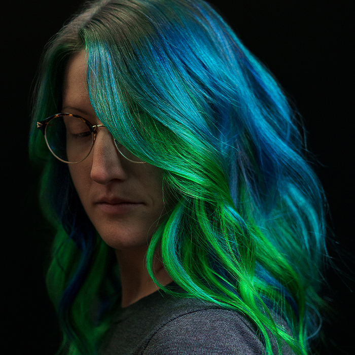 Young woman with a dark shadow root melting into neon blue hair with a vibrant neon green money piece framing the face, created using Danger Jones hair colour.
